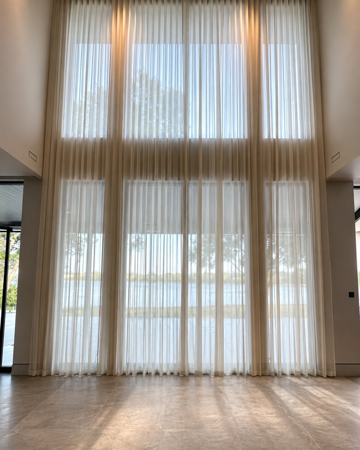 Double-height sheer wave curtains on a ceiling track in front of a lake view
