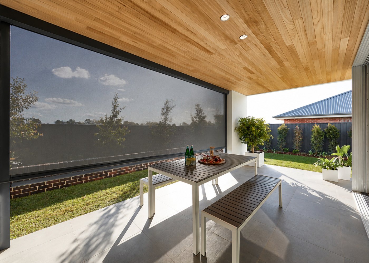 A covered patio with a heavy-duty mesh outdoor blind lowered, dining table set, garden visible through the screen