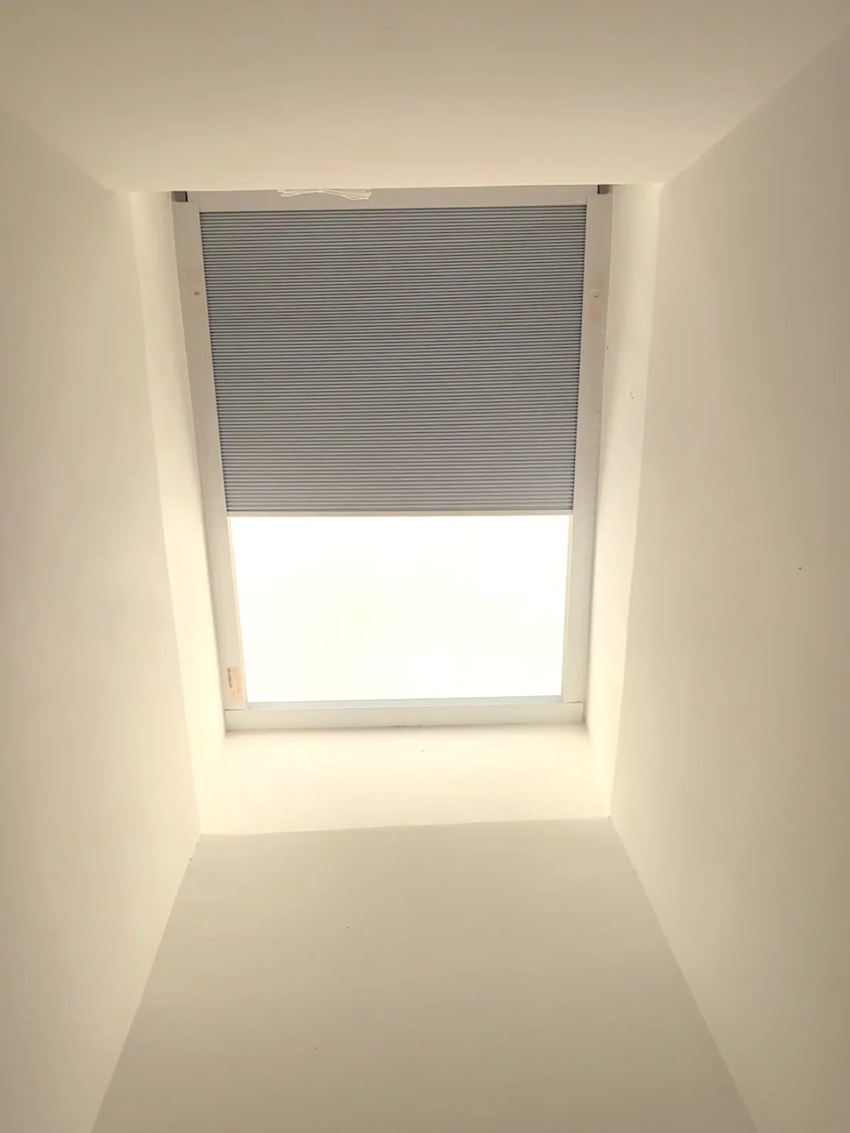 A motorized cellular skylight shade half-drawn across a ceiling skylight in a deep light well